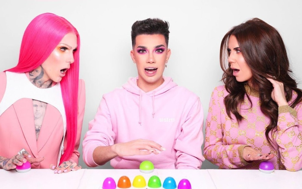 A screenshot of a video featuring Jeffree Star on the left, James Charles in the middle, and Tati Westbrook on the right. They are seated at a white table, all wearing pink clothes, and Jeffree and Tati are both looking at James with shocked expressions.